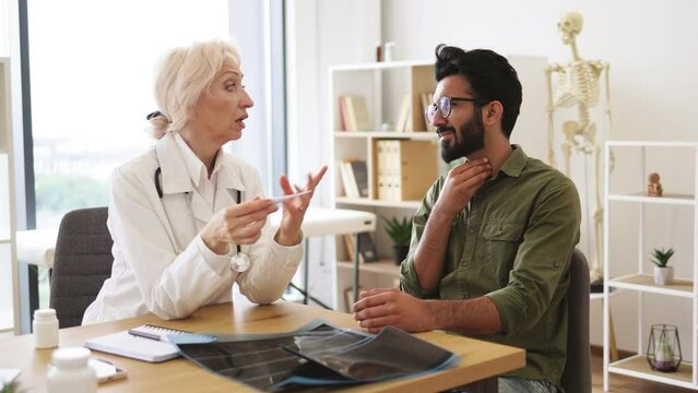 Caucasian senior female doctor and young male with sore throat sitting at table of modern medical center and measuring temperature.
