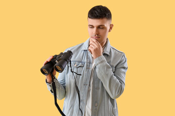 Thoughtful young man with binoculars on yellow background - obrazy, fototapety, plakaty