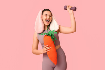 Happy female fitness coach in bunny ears with toy carrot and dumbbell on pink background. Easter...