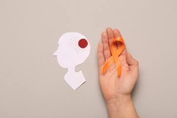 Female hand with orange ribbon and paper human head on grey background. Multiple Sclerosis...