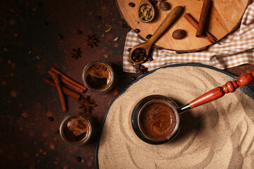 Pot with coffee in sand, cups and spices on dark background