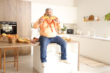 Overweight mature man sitting on table full of unhealthy food in kitchen. Overeating concept