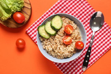 Bowl with tasty oatmeal, tomatoes and cucumber on orange background