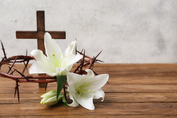 Crown of thorns with white lilies and cross on wooden table near light wall