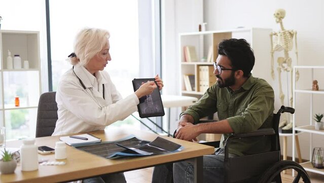 Portrait of senior female doctor and bearded male patient with disability posing at office desk in clinic. Health professional giving recommendation about treatment due to CT scan images.