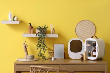 Open cosmetic refrigerator with products, mirror, hand figure and houseplant on table near yellow...