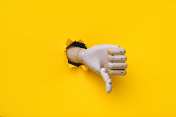 A wooden hand protrudes from a torn hole in yellow paper and shows a thumbs down (dislike). The concept of disapproval and that things will go badly. Artificial intelligence, robot.