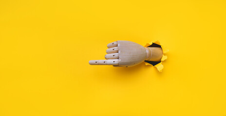A wooden hand protrudes from a torn hole in yellow paper and points its index finger to the left. Concept of direction, adjustment and assistance. Mannequin, robot - obrazy, fototapety, plakaty