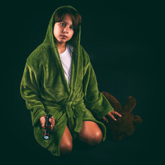 Frightened young boy in a robe kneeling on the floor in the dark with a flashlight and his teddy...
