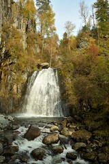 A wide stream of a rapid waterfall flows from a high mountain into a valley yellowed by autumn on a sunny day.