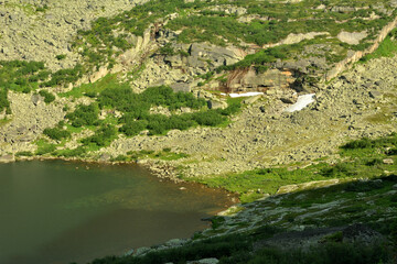 Top view of a fragment of a large lake surrounded by gentle slopes of high mountains with loose stones and sparse thickets of grass.