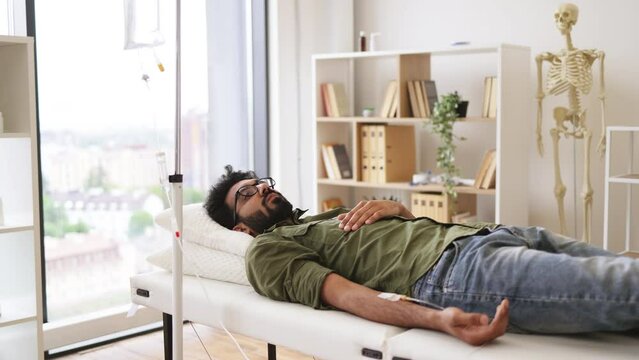 Tired sick male in casual wear drips medicine intravenously while lying on modern light ward. Young bearded man taking dropper with solution for chemotherapy lying on exam couch.