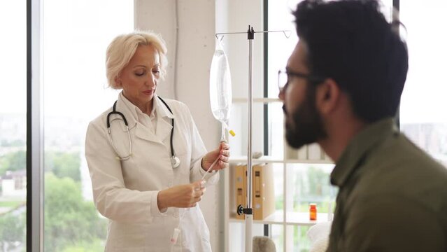 Medical specialist checking drug solution before dripping medicine of patient in exam room. Bearded young man receiving chemotherapy looking at nurse in lab coat preparing a dropper.