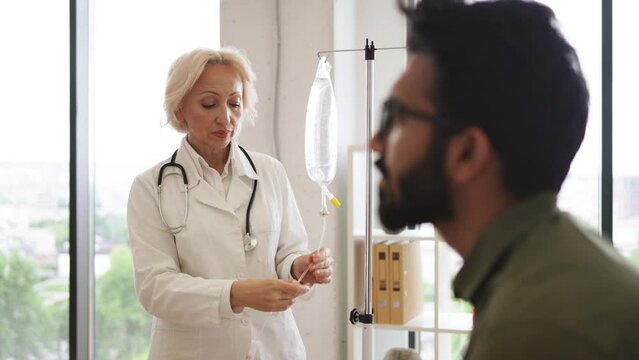 Elderly woman in lab coat testing dropper with solution for chemotherapy near young man sitting on couch in exam room. Healthcare practitioner consulting male patient with cancer in clinic.