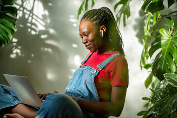 Pleased african american woman with laptop on crossed legs smiling look at screen watching video in earphones sits in room with plants. Satisfied black girl scrolling internet social media on computer - obrazy, fototapety, plakaty