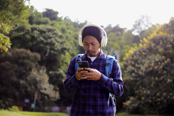 Young Asian man in a casual outfit, sporting a beanie, plaid shirt, and headphones, checks his phone with a puzzled expression on his morning nature walk. A backpack rests comfortably on his shoulder.