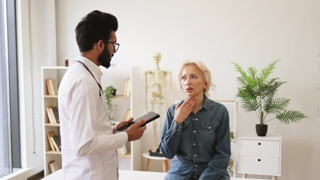 Old woman describing symptoms of illness to young male doctor at moderm medical office. Adult bearded physician giving recommendations and using tablet for writing electronic prescription.