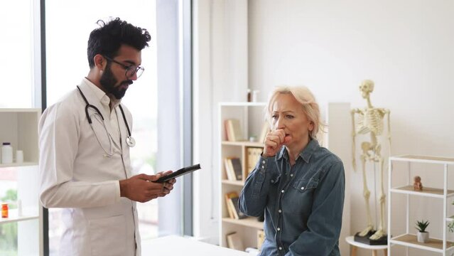 Old woman describing symptoms of illness to young male doctor at moderm medical office. Adult bearded physician giving recommendations and using tablet for writing electronic prescription.