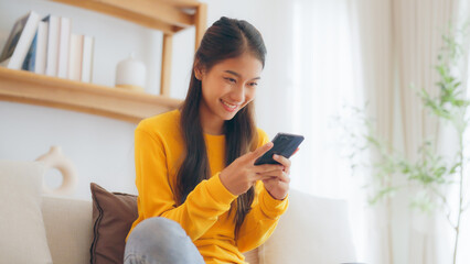Happy young asian woman relax on comfortable couch at home, typing chat message on smartphone,...