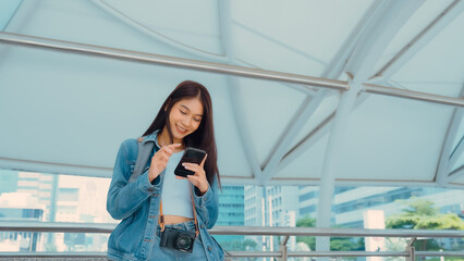 Young asian woman smiling using mobile smart phone outdoor. Happy female tourist wearing jeans jacket and holding smartphone at public - obrazy, fototapety, plakaty