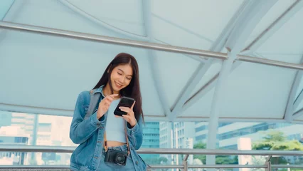 Foto op Canvas Young asian woman smiling using mobile smart phone outdoor. Happy female tourist wearing jeans jacket and holding smartphone at public © Monster Ztudio