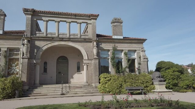 The Epinal Roman house is sublimated in the intricate details of the large arches and imposing columns. Sunlight bathes the building, casting soft shadows and highlighting the surrounding garden.