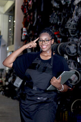 Fototapeta na wymiar Portrait of curly hair African American woman with apron holds laptop computer while stands in thinking pose at auto spare parts store warehouse workplace, surrounded by secondhand engine parts.