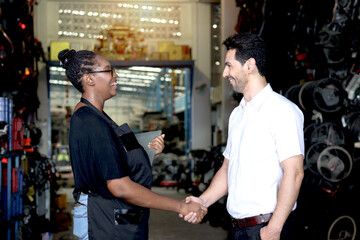 Happy harmony people at workplace, smiling guy making handshake with partner African American woman who working together at auto spare parts store warehouse, surrounded by secondhand engine parts.