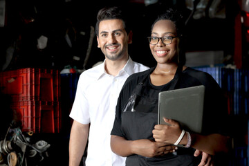 Portrait of happy harmony people working together at workplace, smiling African American woman holds laptop with her colleague at auto spare parts store warehouse, surrounded by secondhand engine part