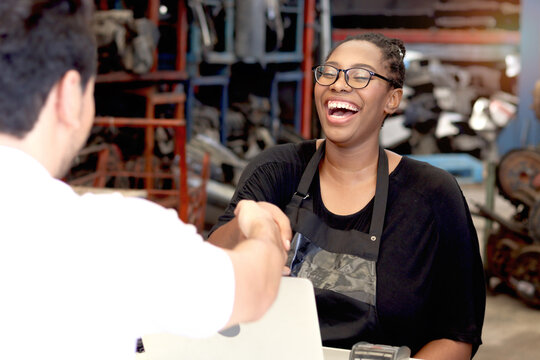 Happy African American woman staff with apron making handshake with customer after completing business deal at auto spare parts store warehouse with secondhand engine parts. Owner offer deal to buyer.