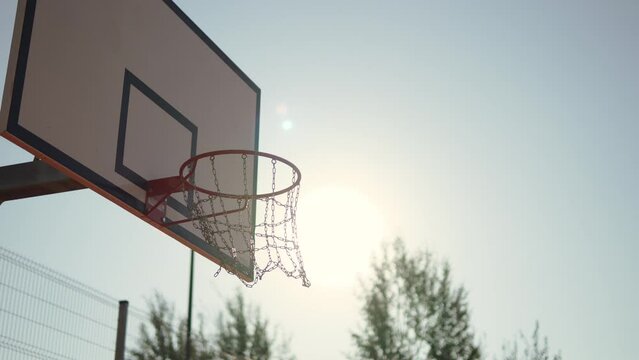 Basketball ball hits hoop in sunlight and causes chains to swing, arc shot. Hit target and earn points on street basketball court. Playing sports and achieving goal in competition outdoor.