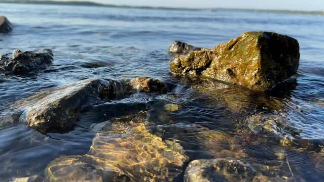 Horizontal video of serene aquatic scene with sunlight dancing on rippling water surface near several rocks. For blogs and articles about environment, cleanliness of water bodies, swimming