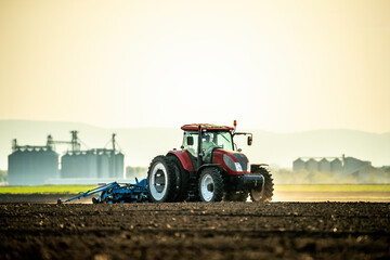 Farmer operates a red tractor, seeding the field against the backdrop of a setting sun - obrazy, fototapety, plakaty