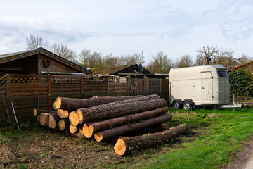 A pile of sawn tree trunks stacked near a fence and a car trailer for transporting animals.