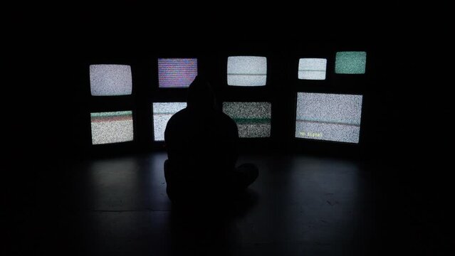 An anxious and isolated person sits in front of a wall of white noise filled televisions media screens in a dark room.