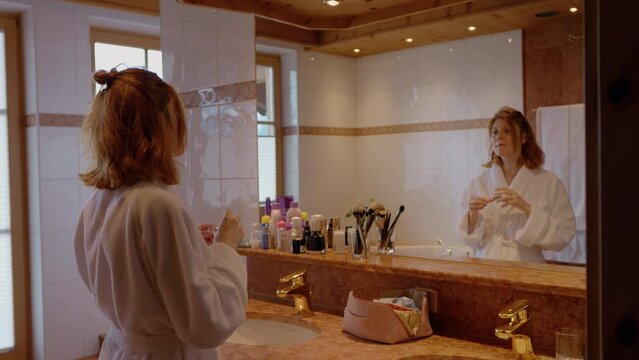 Middle aged woman doing makeup applying lip-gloss looking in mirror. Morning routines of a busy female in the bathroom.