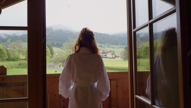Early morning woman standing in white robe on hotel balcony overlooking mountains in the distance. Female looking outside while on vacation.
