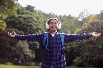 Young Asian man in a casual outfit, sporting a beanie, plaid shirt, backpack, and headphones, stretches his arms wide with a smile, seemingly enjoying the fresh morning air on a nature walk.