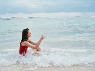 Woman with a beautiful tan tourist in a red swimsuit sitting on the sand on the beach in the ocean in the waves pensive