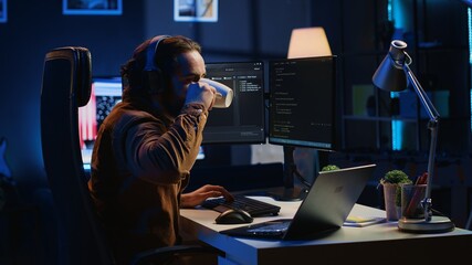 Programmer does multitasking to finish developing PC code, drinks coffee, listens music. IT admin wearing headphones, enjoying hot beverage, solving database errors on computer and laptop, camera A