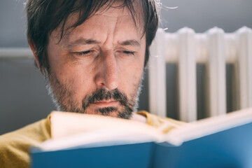 Adult caucasian man with beard reading blue hardcover book in home living room in morning