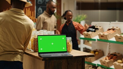 Laptop on stall at supermarket showing greenscreen template, people shopping for organic homegrown products in zero waste eco store. Portable pc with blank chromakey and copyspace. Camera 1.