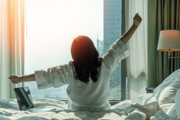 Work-life balance lifestyle quality with happy hotel relaxation on lazy day with Asian woman waking up from good sleep on bed in weekend morning resting in comfort bedroom looking toward city view