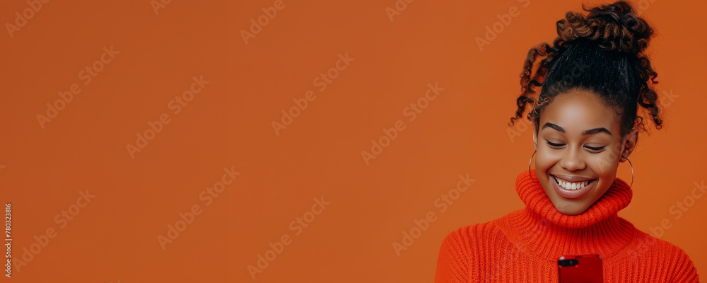 Wall mural Portrait of smiling african american woman using smartphone isolated on orange background, wearing red sweater with copy space for your text message or promotion banner. - Wall murals