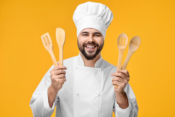 Happy young chef in uniform holding wooden utensils on orange background