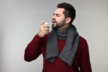 Young man with scarf using throat spray on grey background