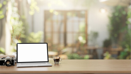 A laptop computer mockup is placed on a wooden table in a green comfortable cafe on a sunny day.
