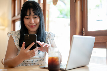 A happy Asian woman sits in a beautiful comfortable coffee shop, using her smartphone.