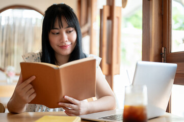 A relaxed and positive young Asian woman reading a book in a beautiful coffee shop on a bright day.