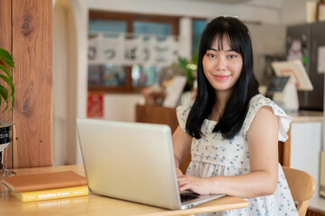A cute Asian woman sits at a table in a coffee shop with her laptop computer, smiling at the camera.
